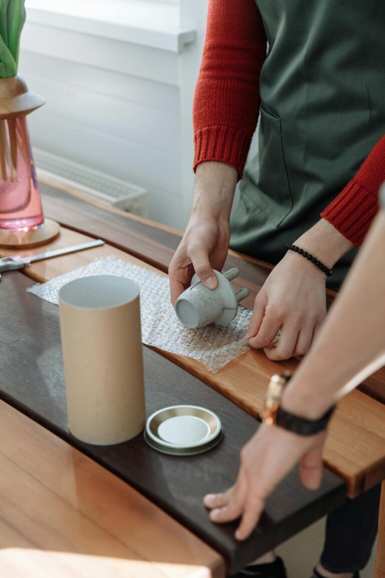 A person carefully wraps a handmade ceramic mug in bubble wrap on a wooden table.