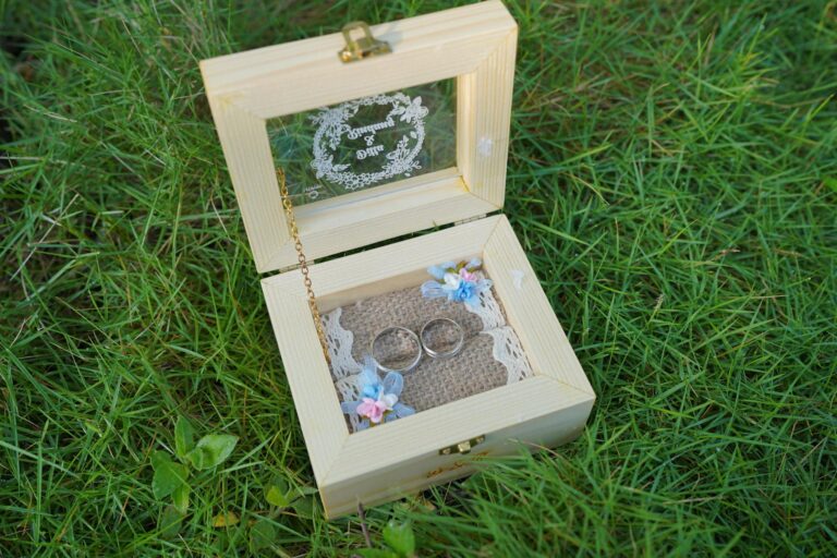 A rustic wooden wedding ring box with engraved glass and wedding bands, placed on grass.